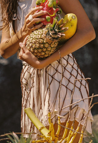 A woman sits on a sandy beach wearing a white short-sleeve top, a white skirt with a brown net pattern, and layered necklaces. Coconuts are visible in the foreground, and tropical foliage and rocks are in the background.