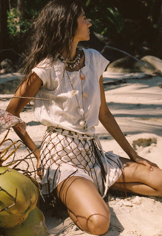 A woman sits on a sandy beach wearing a white short-sleeve top, a white skirt with a brown net pattern, and layered necklaces. Coconuts are visible in the foreground, and tropical foliage and rocks are in the background.