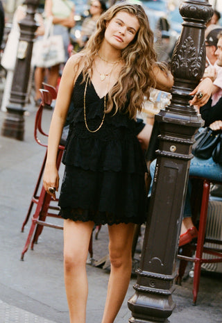 A woman wearing a black tiered ruffle mini dress and gold necklaces stands on a city street next to a dark metal post.