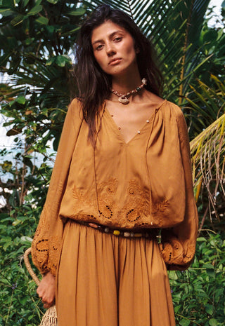 A woman wearing a bohemian-style mustard yellow embroidered top and matching pleated skirt, standing outdoors in front of lush green tropical foliage.