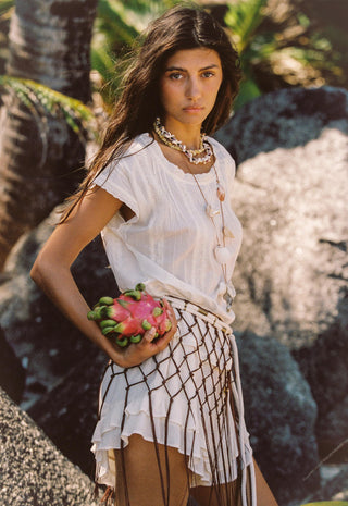 A woman outdoors wearing a white short-sleeve top and a white skirt with a black geometric lattice pattern, holding a pink dragon fruit. She is wearing layered gold necklaces.