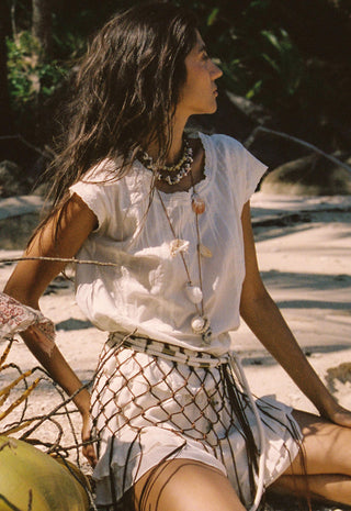 A woman sitting on a sandy beach wearing a white short-sleeve top, a white skirt with a black fishnet overlay, and layered bohemian necklaces.