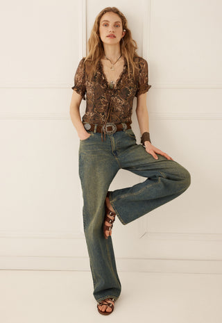 A woman modeling a brown patterned short-sleeve blouse and wide-leg blue jeans with brown sandals, posing against a white paneled wall.