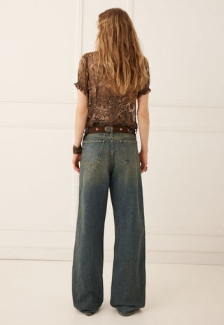 A woman viewed from behind wearing a brown patterned short-sleeve top, a brown belt, and wide-leg blue jeans against a white paneled wall.