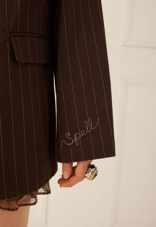 Close-up of a dark brown pinstripe blazer with thin white vertical stripes, featuring a cursive embroidered 'Spell' logo on the cuff and a hand holding a ring near the hem.