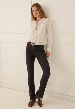 A woman modeling a white long-sleeve lace blouse tucked into dark charcoal jeans with a brown belt and brown sandals, standing against a white paneled wall.