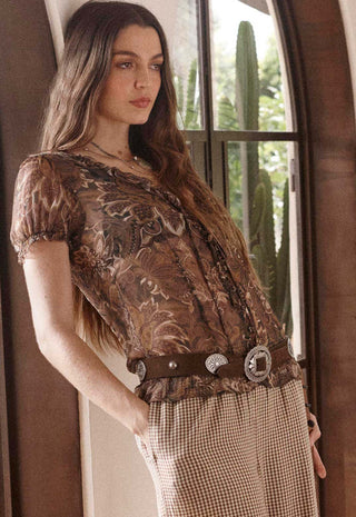 A woman wearing a brown floral lace top and a brown patterned skirt, accessorized with a wide brown belt featuring silver conchos, standing in front of a window with cacti visible outside.