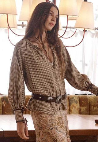 A woman wearing a brown and beige gingham long-sleeve blouse with a dark brown belt and a matching patterned skirt, standing in a warmly lit indoor setting with a chandelier and upholstered bench in the background.