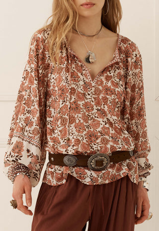 A woman wearing a long-sleeve bohemian-style blouse with a rust and brown floral paisley print on a cream background, styled with a dark brown belt and brown pants.