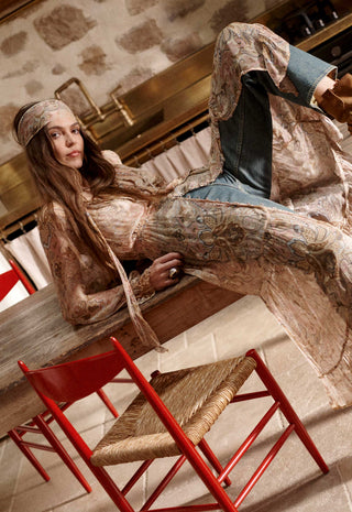 a-woman-reclines-on-a-wooden-table-in-a-rustic-interior-wearing-a-long-sleeve-bohemian-style-patterned-dress-in-beige-brown-and-blue-tones-with-blue-jeans-red-chairs-are-visible-in-the-foreground