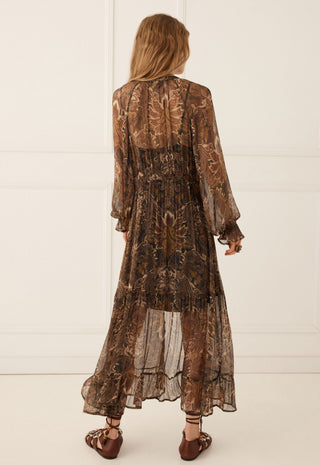 A woman viewed from behind wearing a sheer brown patterned midi dress with long sleeves and ruffled cuffs, paired with brown strappy sandals, set against a white paneled wall.