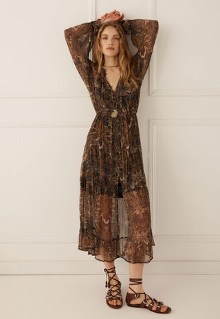 A woman modeling a long-sleeved brown and black floral print midi dress with a tie-front waist and a V-neckline, paired with brown strappy flat sandals, standing against a white paneled wall.