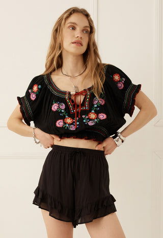 A woman modeling a black short-sleeve crop top with colorful floral embroidery and a red tie detail, paired with black ruffled shorts against a white paneled wall.