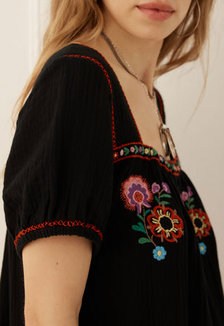 Close-up of a woman wearing a black short-sleeve top with colorful floral embroidery and red contrast stitching on the neckline and sleeve cuffs.