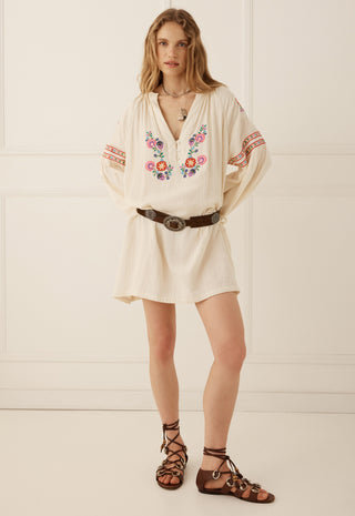 A woman modeling a cream-colored bohemian mini dress with colorful floral embroidery on the chest and sleeves, styled with a brown belt and brown lace-up sandals against a cream paneled wall.