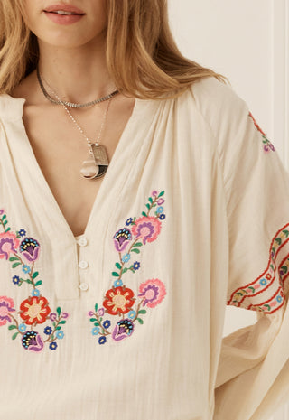 A close-up of a woman wearing a cream-colored bohemian blouse with colorful floral embroidery on the front and sleeves, accessorized with layered silver necklaces.