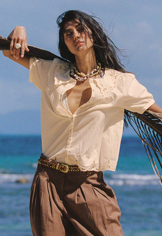a-woman-stands-outdoors-by-the-ocean-wearing-a-loose-white-embroidered-blouse-brown-pleated-pants-and-a-gold-belt-with-a-palm-frond-draped-over-her-shoulder