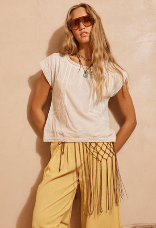 A woman wearing a white short-sleeve top, a pair of yellow pants with a brown net patterned sarong tied around her hips. Against a brown background.