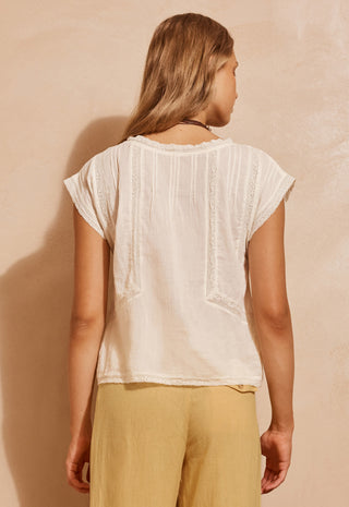 A woman shown from the back wearing a cream-colored short-sleeve top with vertical textured stripes and lace-like detailing, paired with mustard yellow pants, set against a textured beige wall.