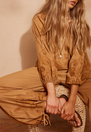 A woman wearing a matching tan embroidered blouse and wide-leg pants, seated on a woven stool against a warm beige wall.