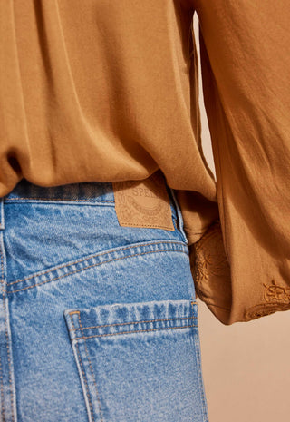 Close-up of a person wearing a camel-colored long-sleeve blouse tucked into light-wash blue jeans, showing the back waistband and a small tan brand patch.