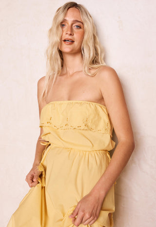 a-blonde-woman-wearing-a-yellow-strapless-midi-dress-with-a-ruffled-eyelet-bodice-standing-against-a-textured-off-white-wall