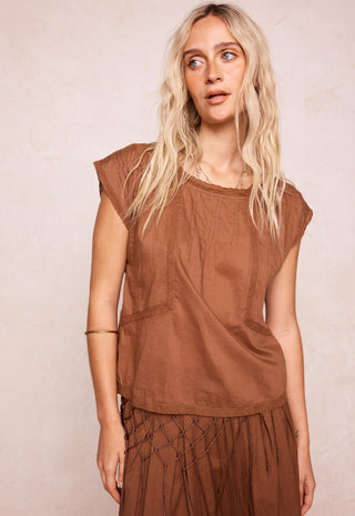 A woman with long blonde hair wearing a brown sleeveless top and matching brown skirt, standing against a textured light beige background.