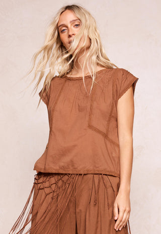 A woman modeling a brown short-sleeve top and matching brown fringed skirt against a light beige textured background.
