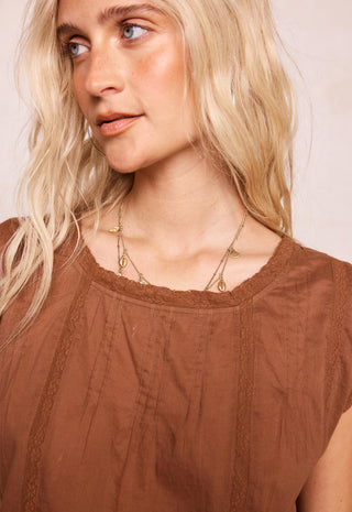 A close-up of a blonde woman wearing a brown textured top and layered gold necklaces, looking off-camera against a light beige background.