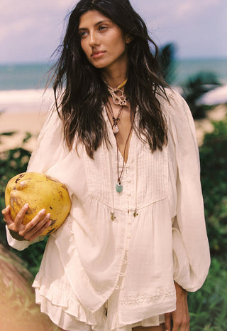 A woman on a beach wearing a loose white bohemian-style blouse and holding a yellow coconut, with layered necklaces and a tropical ocean background.