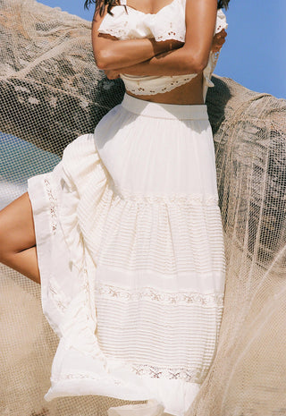 A woman wearing a white embroidered maxi skirt and matching white crop top, posed outdoors against a blue sky with sheer beige fabric draped around her.