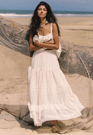 A woman stands on a sandy beach wearing a long white tiered maxi dress with ruffled trim. A large beige fishing net lies behind her, with the ocean and sky in the background.