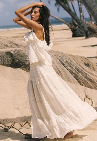 A woman wearing a long white tiered boho maxi dress stands on a sandy beach next to a large driftwood log, with palm trees and the ocean in the background.