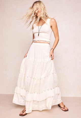 A woman modeling a white sleeveless crop top and a long white tiered skirt with embroidered details, paired with brown sandals, against a neutral beige studio backdrop.