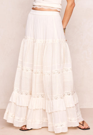 A long tiered white maxi skirt with delicate lace trim and horizontal textured bands, shown on a model against a light beige textured wall.
