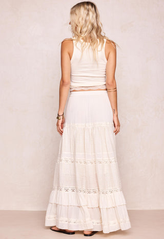 A woman shown from the back wearing a sleeveless cream top and a long cream tiered skirt with lace trim, standing against a light beige textured wall.