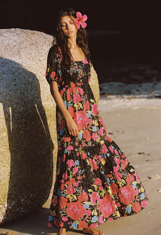 A woman stands barefoot on a sandy beach beside a large rock, wearing a black maxi dress with a vibrant pink, red, and blue floral print. She has a pink flower tucked into her dark hair.