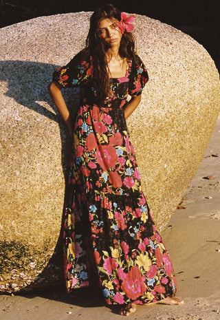 A woman stands barefoot outdoors against a large rock, wearing a long black maxi dress with a vibrant pink, red, and blue floral print and a pink flower in her hair.