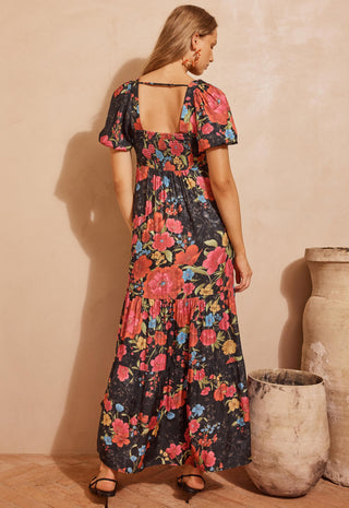 A woman wearing a black maxi dress with a vibrant pink, red, and blue floral print, shown from the back against a warm beige textured wall with rustic clay pots.