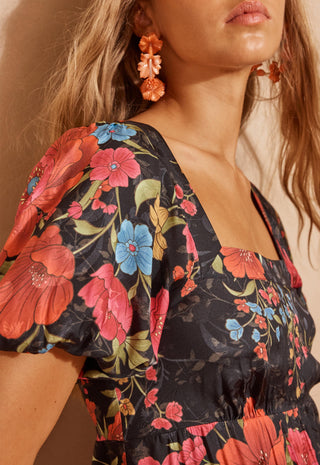 A woman modeling a black maxi dress with a vibrant red, pink, and blue floral print, standing against a textured beige wall.