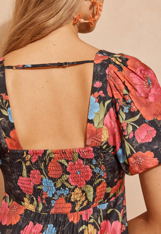 A back shot of a woman modeling a black maxi dress with a vibrant red, pink, and blue floral print, standing against a textured beige wall.