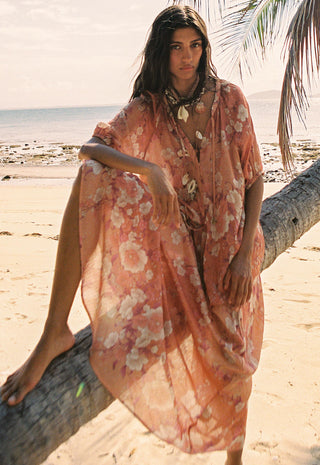 A woman on a sandy beach sits on a fallen palm tree wearing a flowing peach floral kaftan dress. The background shows the ocean, sand, and palm leaves.