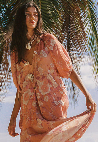A woman wearing a loose, short-sleeve peach floral cover-up dress with a tie neckline, standing outdoors under palm leaves against a bright sky.