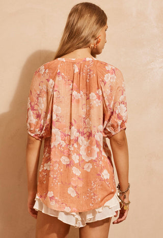 A woman shown from the back wearing a peach-colored short-sleeve blouse with a pink and white floral print, paired with white ruffled shorts, set against a textured beige wall.