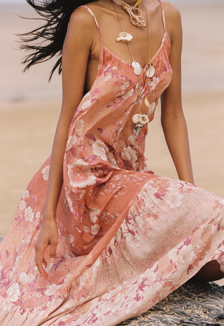 A woman seated on a beach wearing a flowing floral maxi dress in soft peach and pink tones with white flower motifs. The dress has thin straps and a backless open design with shell-like embellishments.
