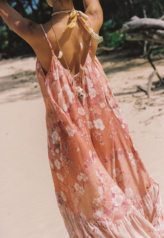 A woman on a sandy beach wearing a peach-pink floral halter maxi dress, shown from behind.