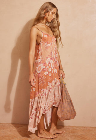 A woman stands barefoot against a warm beige wall, wearing a peach and pink floral spaghetti-strap maxi dress and holding a woven tote bag.