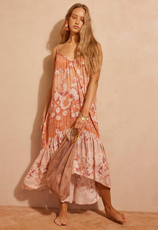 A woman modeling a sleeveless, high-low floral maxi dress in a peach and pink color palette with a tie-dye pattern, standing against a textured beige wall.