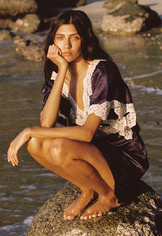 A woman crouches barefoot on a rock by the water, wearing a dark purple satin robe with white lace trim.