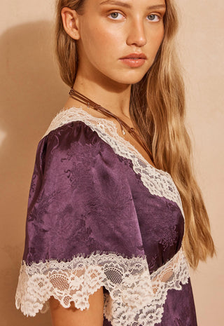 A woman wearing a purple satin slip dress with white lace trim, photographed against a warm beige background.